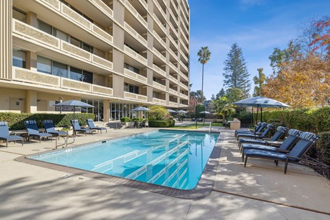 A large swimming pool surrounded by lounge chairs and umbrellas in front of a multi-story apartment building.