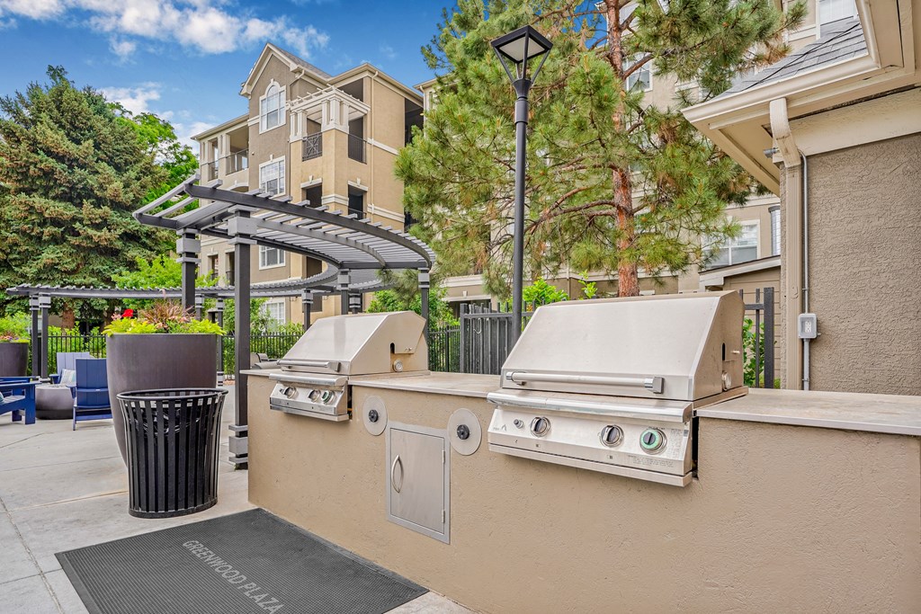 A BBQ grill is on a patio with a trash can and a tree.