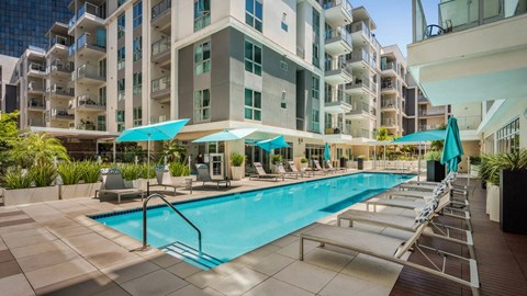A large swimming pool surrounded by lounge chairs and umbrellas in front of a modern apartment building.