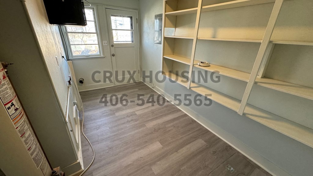 A room with a white door, a window, and a wall with a TV and a shelf.