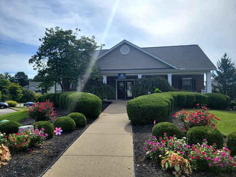 A building with a grey roof and a signboard is surrounded by green bushes and flowers.