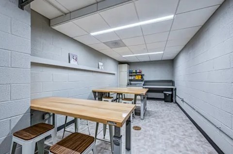 A room with white walls and a grey floor has a long table with chairs and a shelf with items on it.