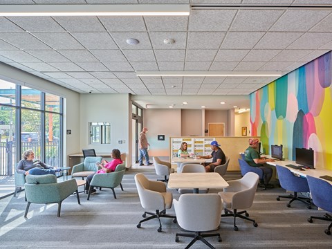 A group of people are sitting and standing in a room with a colorful mural on the wall.