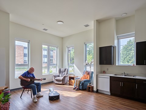 A man is playing guitar in a spacious room with wooden flooring and white walls.