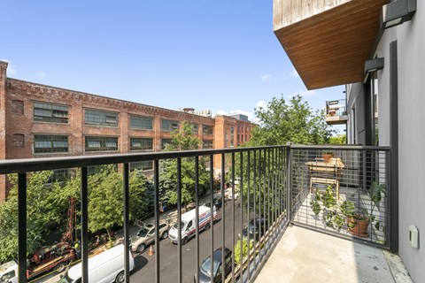 A balcony with a black railing overlooks a parking lot and a brick building.