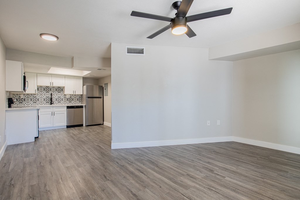 an empty living room with a ceiling fan and a kitchen in the background