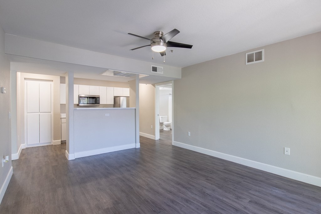 an empty living room with a ceiling fan and a kitchen in the background