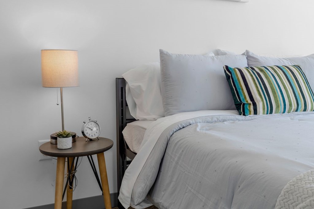 A bed with a grey comforter and striped pillow beside a table with a lamp and a small plant.