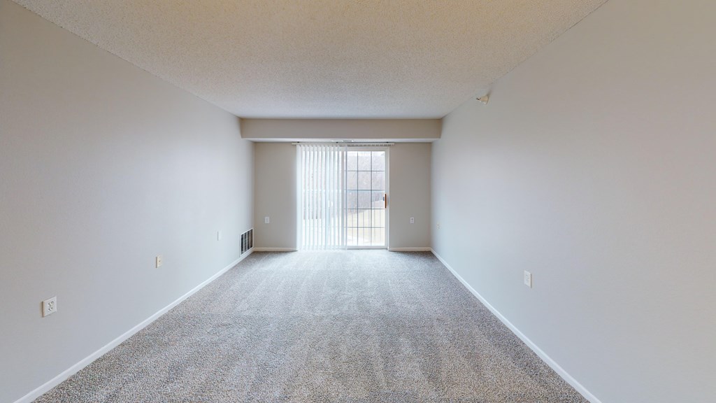 A large, empty room with a carpeted floor and a window with blinds.