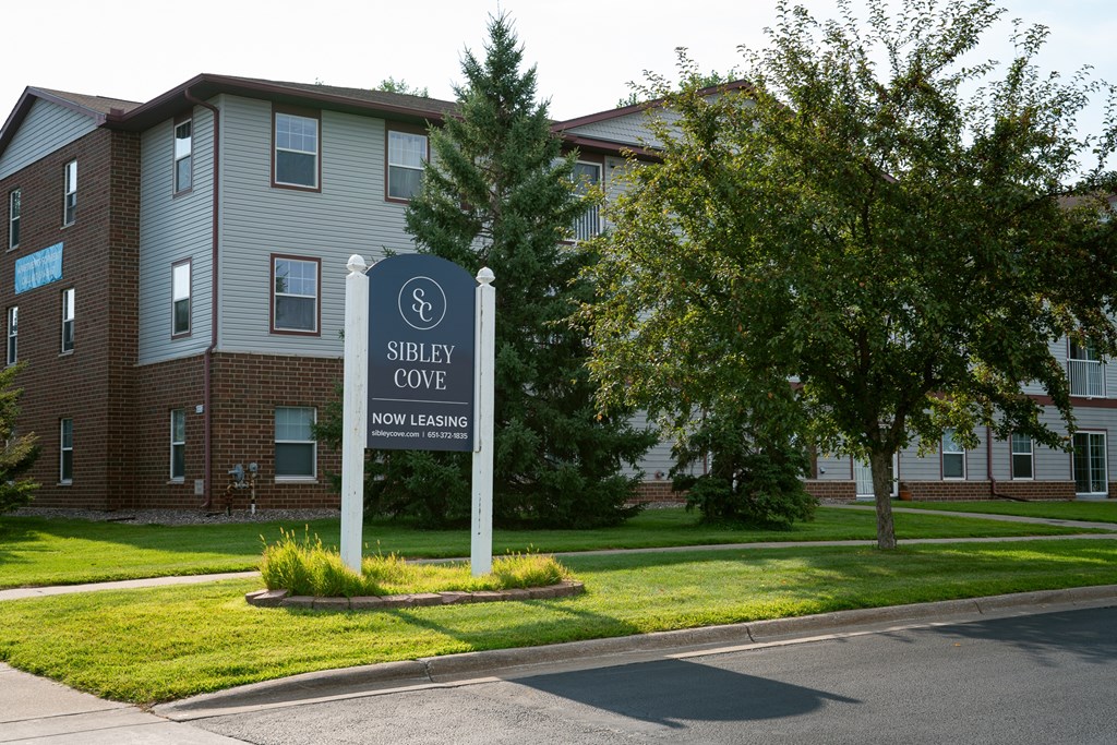 A sign for Sibley Cove, a now leasing apartment complex, stands in front of a brick building.