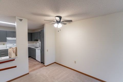 A room with a ceiling fan and a kitchen area in the background.