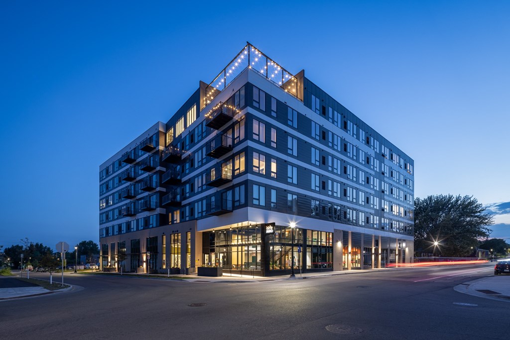 A modern multi-story building with a glass facade is illuminated from within and surrounded by a dark sky.