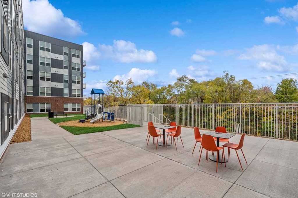A patio with red chairs and a table is surrounded by a fence and a building.