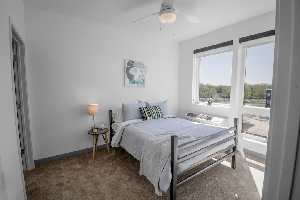 A bedroom with a bed, a nightstand with a lamp, and a window overlooking a parking lot.