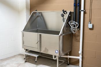 A large industrial machine with a grey top and a white base is situated in a room with a white wall and a brown wall.