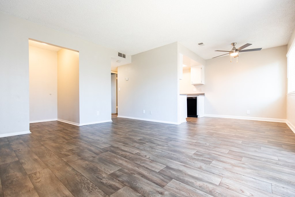 an empty living room with a ceiling fan and a kitchen in the background
