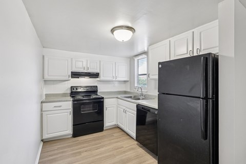 A black refrigerator stands in a kitchen with white cabinets and a wooden floor.