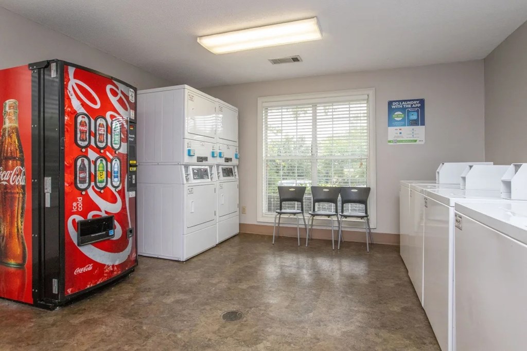 A Coca-Cola vending machine sits in a kitchen with white appliances and a window.