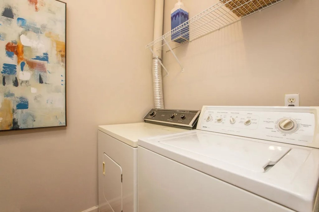 A white washing machine sits in a laundry room with a painting on the wall.