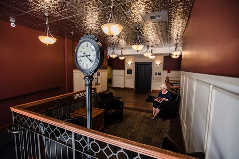 A woman is sitting on a black couch in a room with a large clock on a pole.