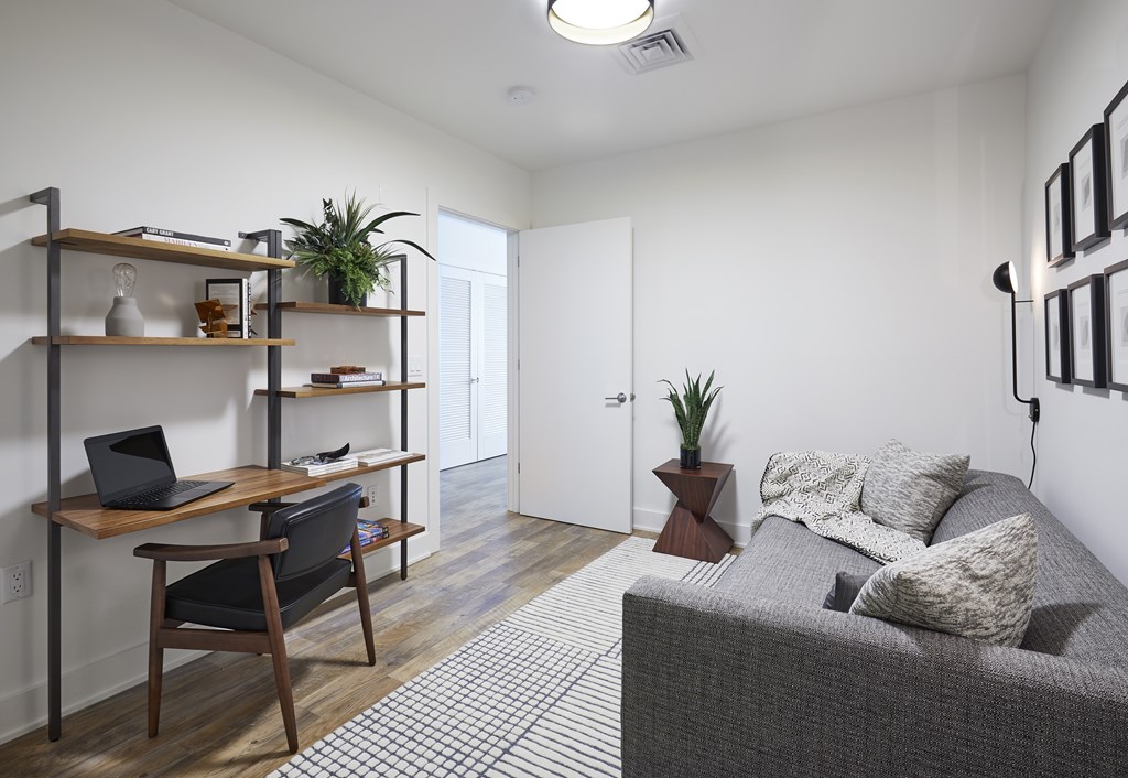 A room with a grey couch, a wooden desk with a laptop, a chair, and a shelf with various items.