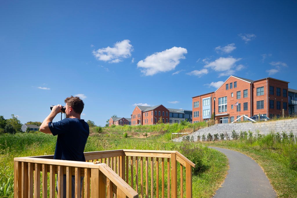 A man is standing on a wooden deck and looking through binoculars at a large red building.