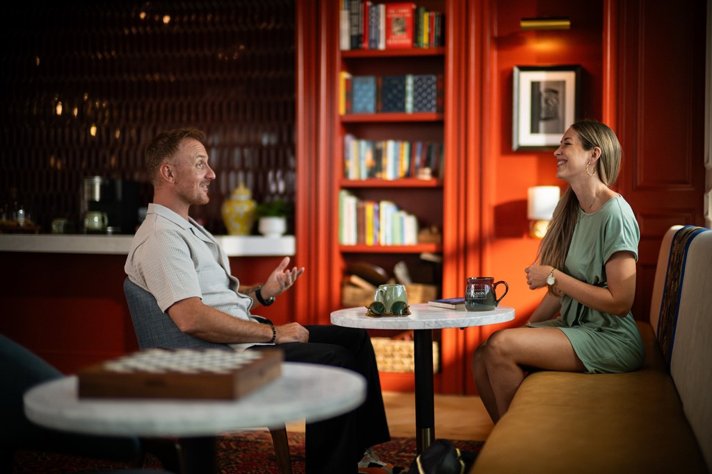 A man and a woman are having a conversation in a cozy room with a bookshelf in the background.