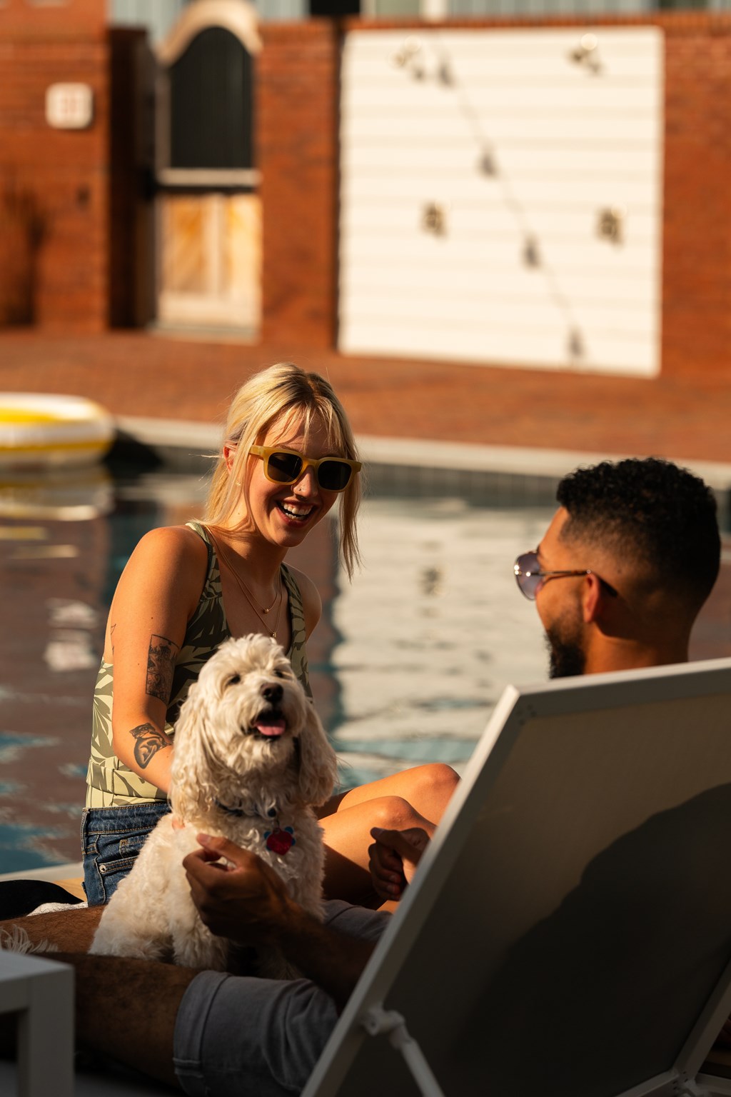 A woman and a man are sitting by a pool with a dog, looking at a laptop.