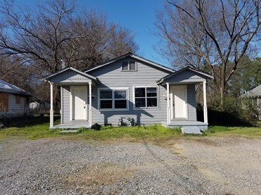 a small gray house with a gravel driveway and trees in the background
