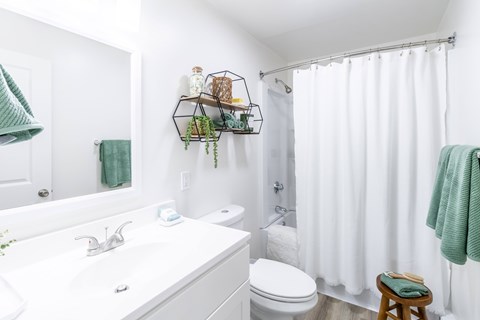 A white bathroom with a white toilet, sink, and a green towel hanging on the shower curtain.
