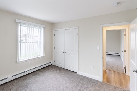 A room with a white door, a window with blinds, and a carpeted floor.