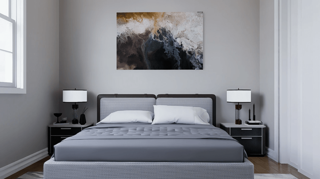 A bed with a grey bedspread and two white pillows is in the foreground of a room with a large abstract painting on the wall.