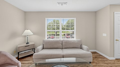 A living room with a grey couch, a lamp, and a window with a view of a parking lot.