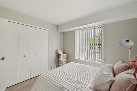 A bedroom with a bed, pillows, a lamp, and a window with blinds.