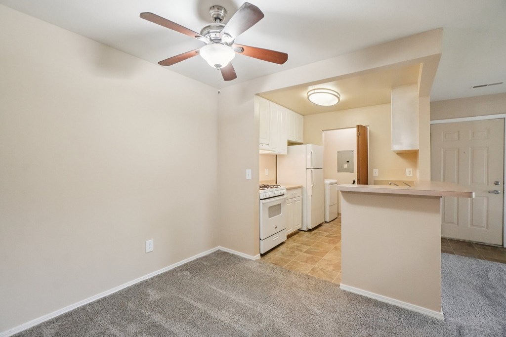 A room with a ceiling fan and a kitchen area in the background.