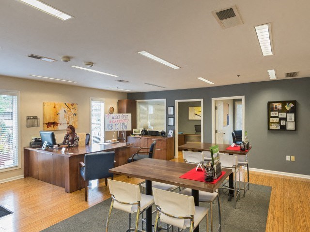 a room with a table and chairs and a woman on a computer at a desk