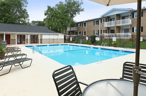 A pool with chairs and a table is surrounded by a fence and apartment buildings.