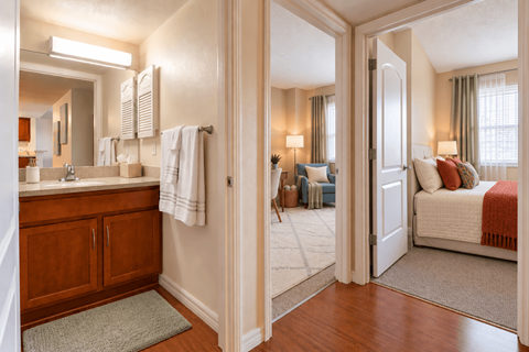 A bathroom with a mirror, sink, and a bedroom with a bed and a window.