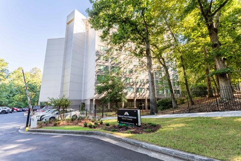 A modern building with a white facade is surrounded by trees and has a black sign in front.
