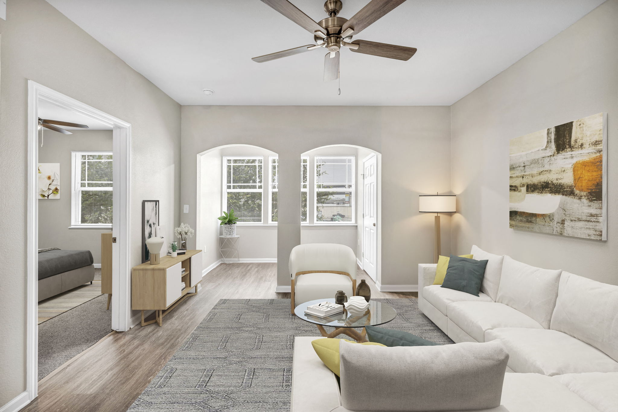 A living room with a grey rug, a white sofa, a grey armchair, a wooden side table, a white coffee table, a white ceiling fan, a white door, a white wall, a white window, a white lamp