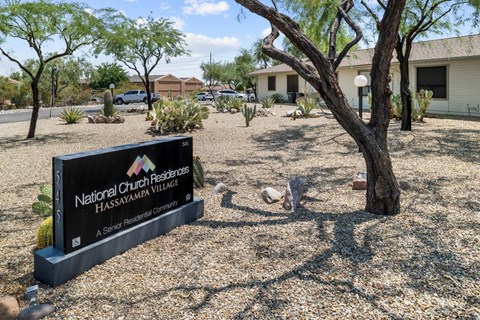 A sign for National Church Residences Hassayampa Village stands in front of a tree.