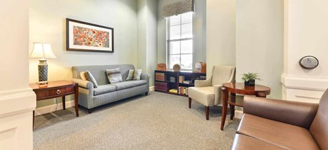 A living room with a grey sofa, a brown leather chair, a wooden side table with a lamp, a small table with a plant, and a framed picture on the wall.