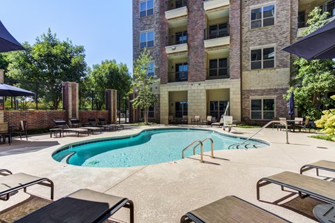 A large swimming pool surrounded by lounge chairs and umbrellas in front of a multi-story apartment building.