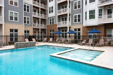 A swimming pool surrounded by chairs and umbrellas in front of apartment buildings.
