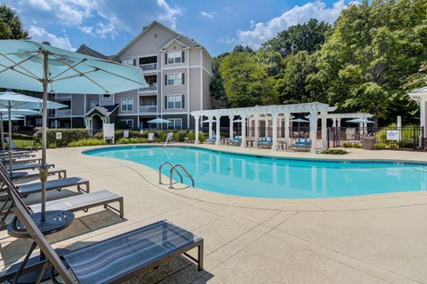 A swimming pool surrounded by sun loungers and umbrellas in front of apartment buildings.