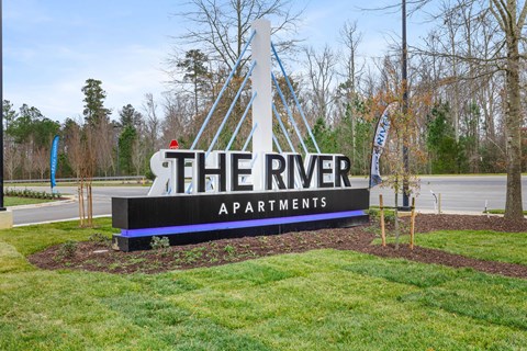 A sign for The River Apartments is displayed in front of a grassy area.