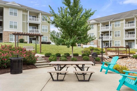 A large tree is in the middle of a courtyard with two picnic tables and two blue chairs.