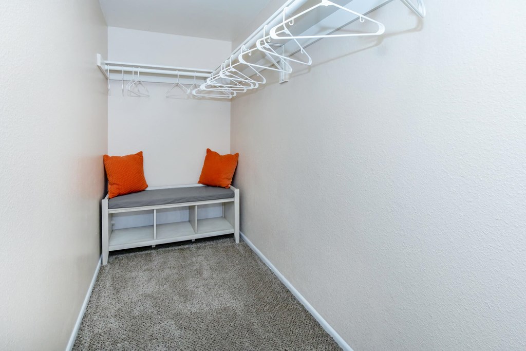 a walk in closet with a bench with orange pillows and white walls