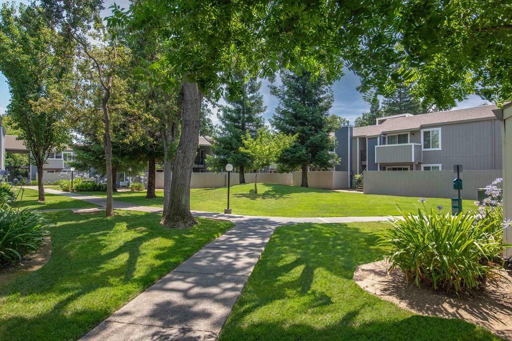 A sidewalk between apartment building with trees and grass and pet station at Willow Grove Apartments, California 