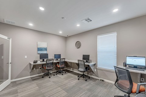 A room with a carpeted floor and a white ceiling with lights, containing a desk with a laptop and a monitor, a chair, and a wall clock.